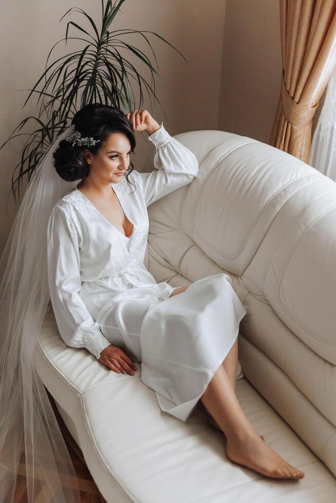 an incredibly beautiful bride in the morning in a white robe and a long veil sits on the bed in her bedroom. The bride poses in the morning before the wedding ceremony. photo