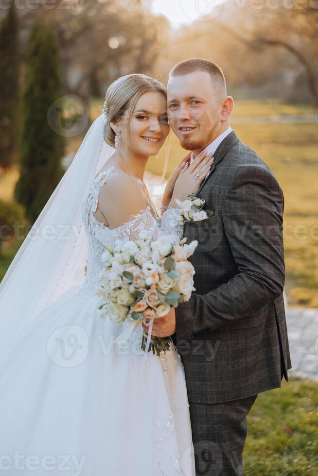 Stylish, young groom and beautiful bride in a long white dress and a long veil with a bouquet in their hands, hugging in the park in the autumn nature. Wedding portrait of newlyweds. photo