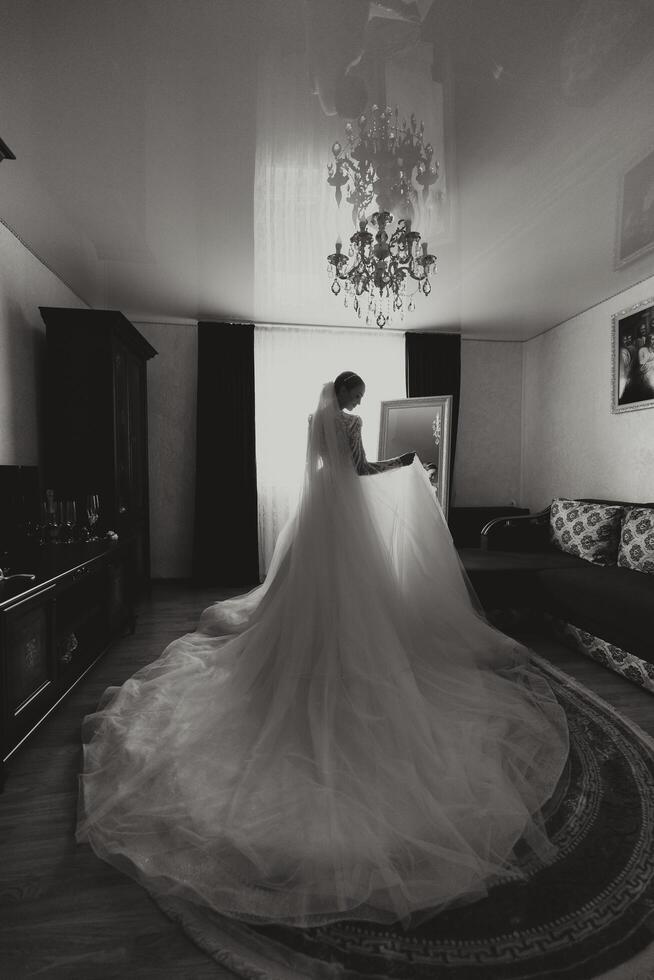 maravilloso contento lujo elegante morena novia cerca un ventana en el antecedentes de Clásico habitación. novia con hermosa manicura participación Boda vestido. Boda preparación de novia. foto