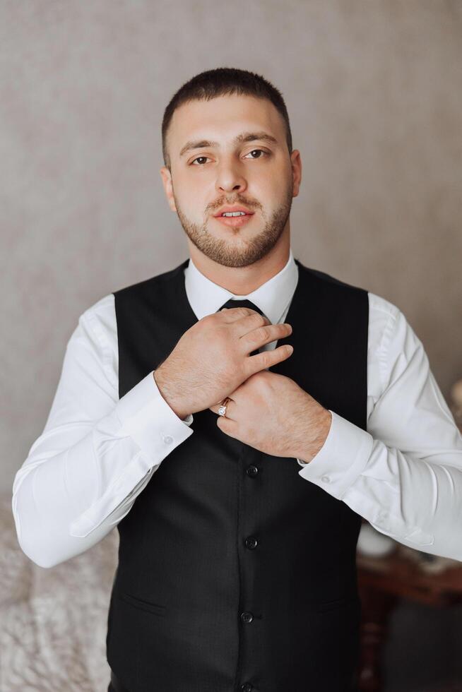a young man in a white shirt stands by the window in the room and puts on a tie. The groom gets dressed in the morning and prepares for the wedding. photo