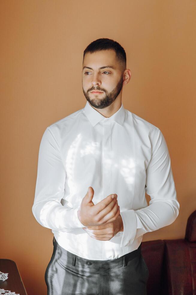 un hombre en un blanco camisa soportes por el ventana en el habitación y sujeta el botones en su collar y mangas reloj en mano. elegante negocio retrato de un hombre, de cerca foto. el novio es preparando. foto