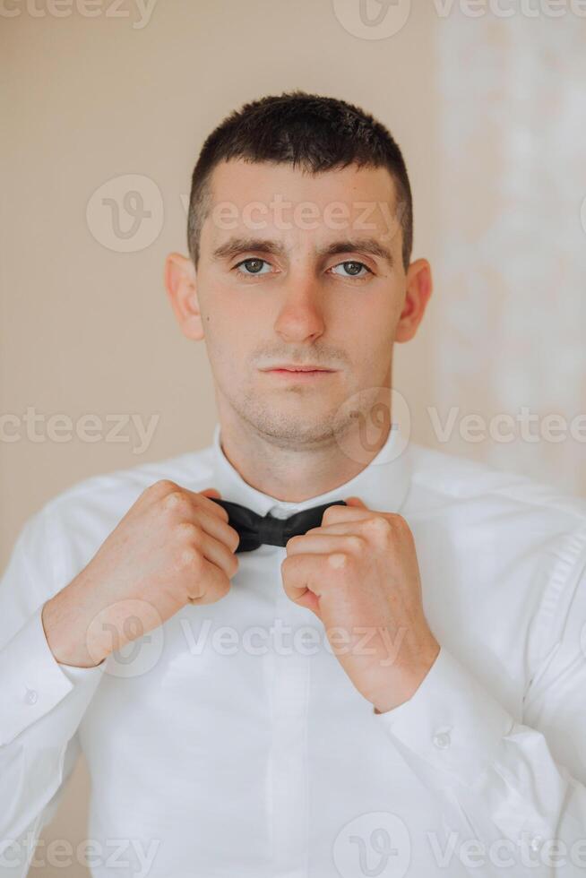 a young man in a white shirt stands by the window in the room and puts on a tie. The groom gets dressed in the morning and prepares for the wedding. photo