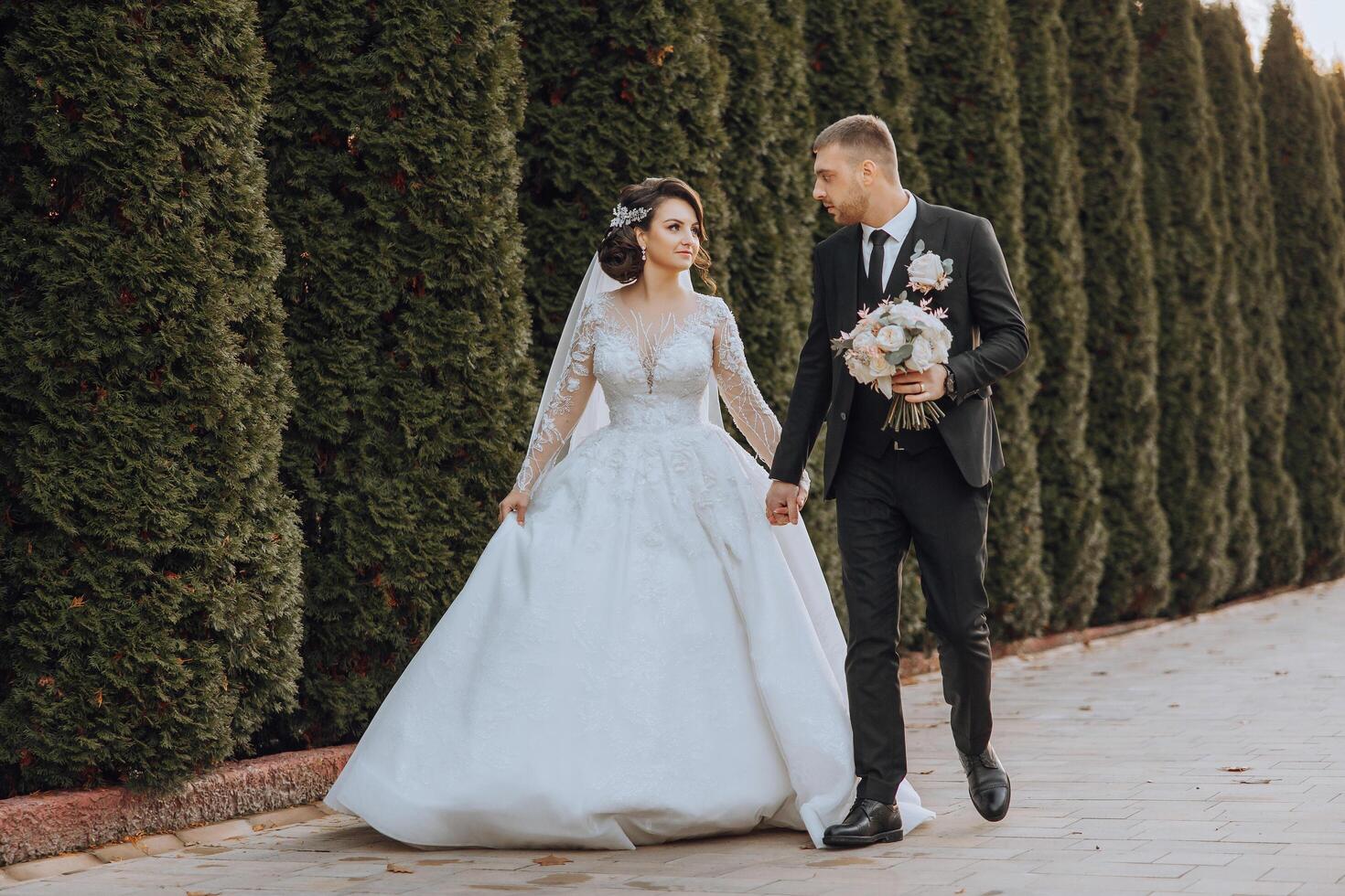 A wedding couple is walking in nature on an autumn day. Happy young bride and elegant groom holding hands. A stylish couple of newlyweds on their wedding day. photo