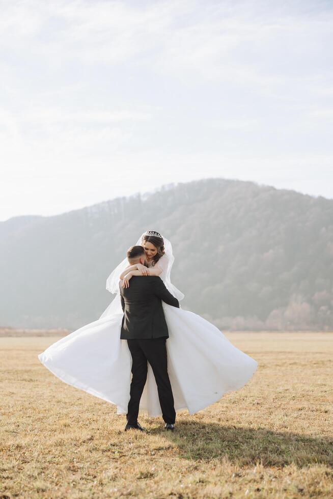 Stylish groom holds beautiful bride in long white dress in his arms and dances on green grass in autumn park, forest under sunlight. Wedding photography, portrait of smiling newlyweds. photo