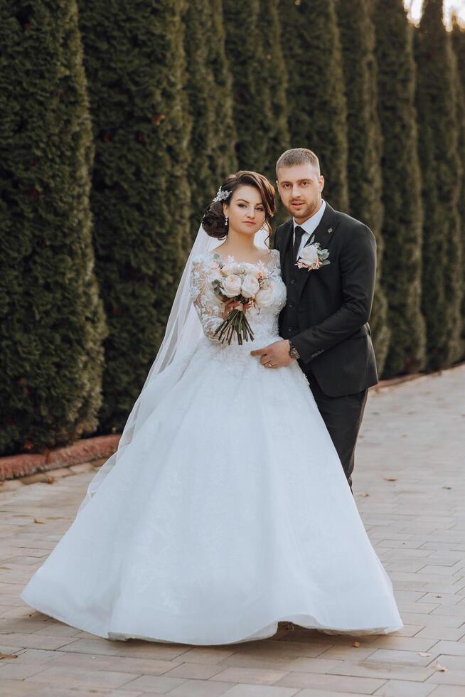 Stylish, young groom and beautiful bride in a long white dress and a long veil with a bouquet in their hands, hugging in the park in the autumn nature. Wedding portrait of newlyweds. photo