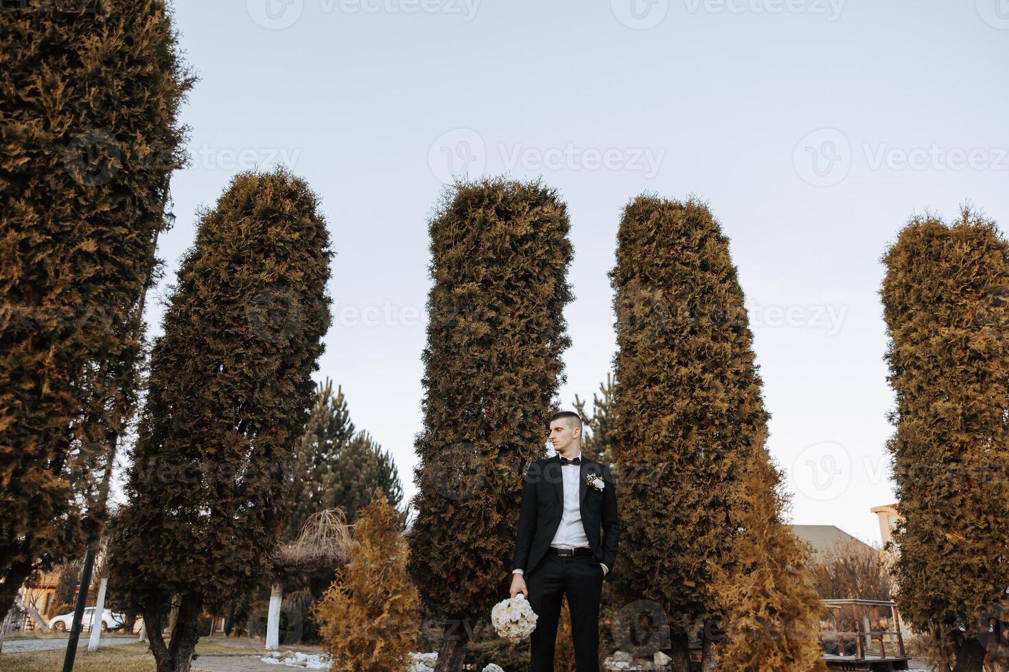 Portrait of a handsome young groom on an autumn day outdoors in an elegant suit and holding a wedding bouquet of flowers. Outdoor photo. Handsome man in a business suit. photo