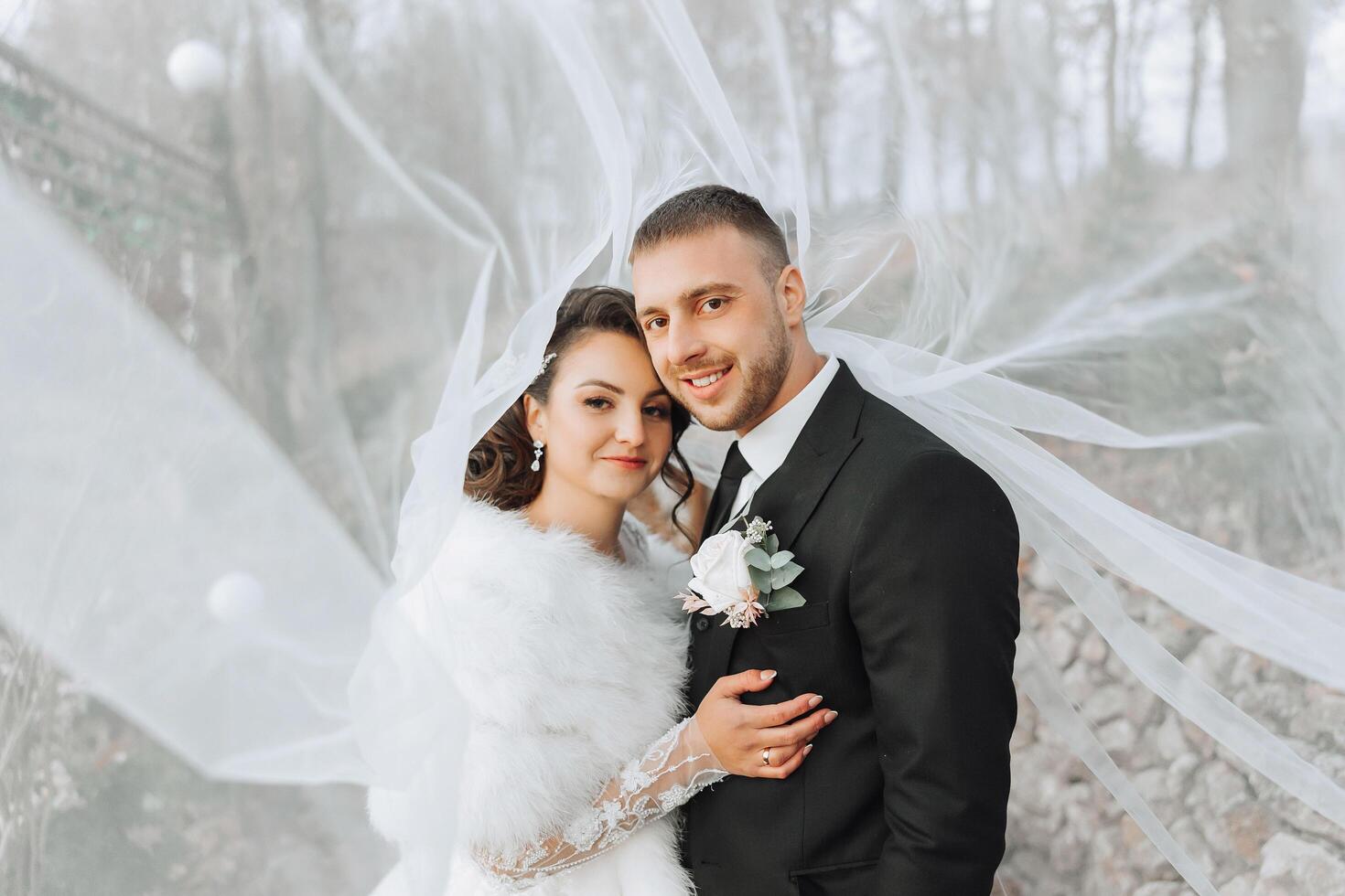 Stylish, young groom and beautiful bride in a long white dress and a long veil with a bouquet in their hands, hugging in the park in the autumn nature. Wedding portrait of newlyweds. photo