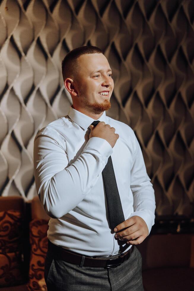 a young man in a white shirt stands by the window in the room and puts on a tie. The groom gets dressed in the morning and prepares for the wedding. photo