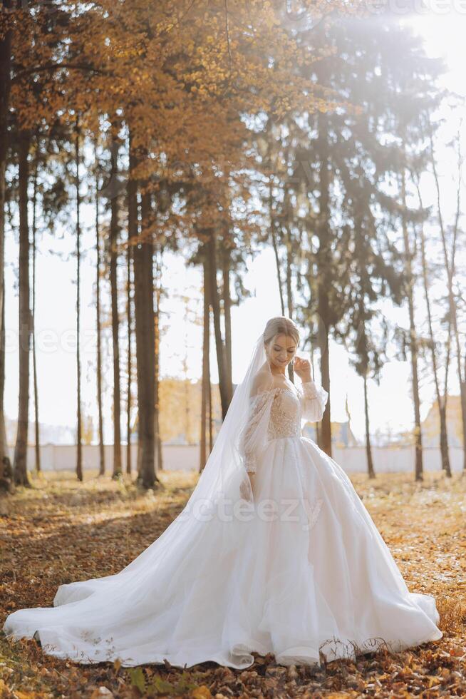 A blonde bride in a white dress with a long train holds the dress and walks down the path covered with autumn leaves. Wedding photo session in nature. Beautiful hair and makeup. Celebration