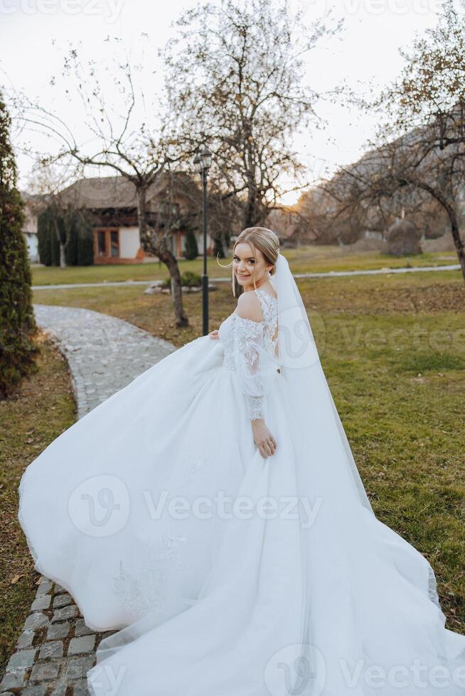 A blonde bride in a white dress with a long train holds the dress and walks along the stone path. Autumn. Wedding photo session in nature. Beautiful hair and makeup. celebration