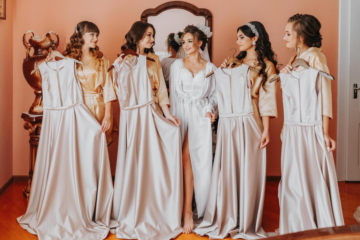 Young bridesmaids in silk robes show off their dresses in a hotel room. Beautiful women celebrating a bachelorette party next to the bride in themed pajamas. photo