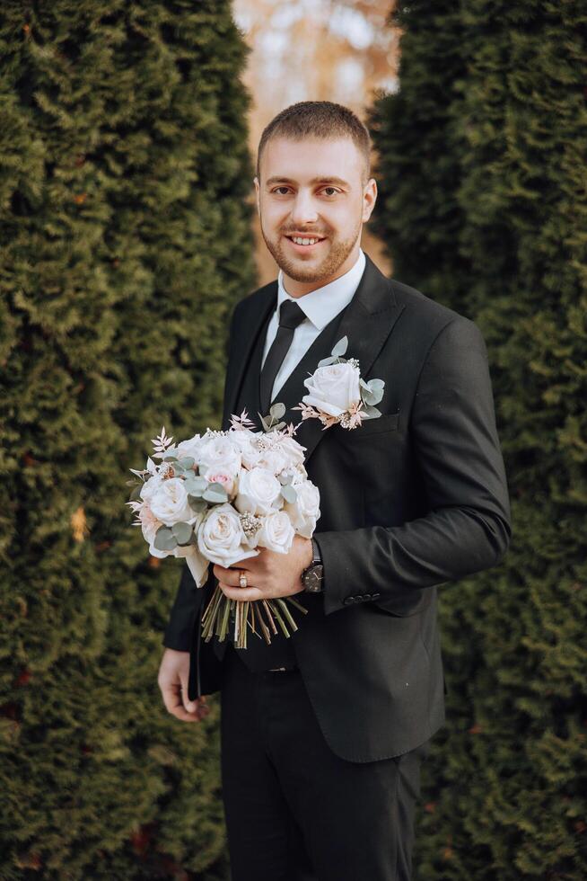 Portrait of a handsome young groom on an autumn day outdoors in an elegant suit and holding a wedding bouquet of flowers. Outdoor photo. Handsome man in a business suit. photo