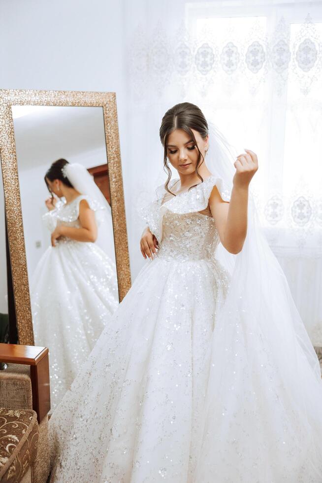 A young European woman with dark hair in a wedding dress stands by a mirror in a studio decorated with flowers. Young beautiful bride. photo