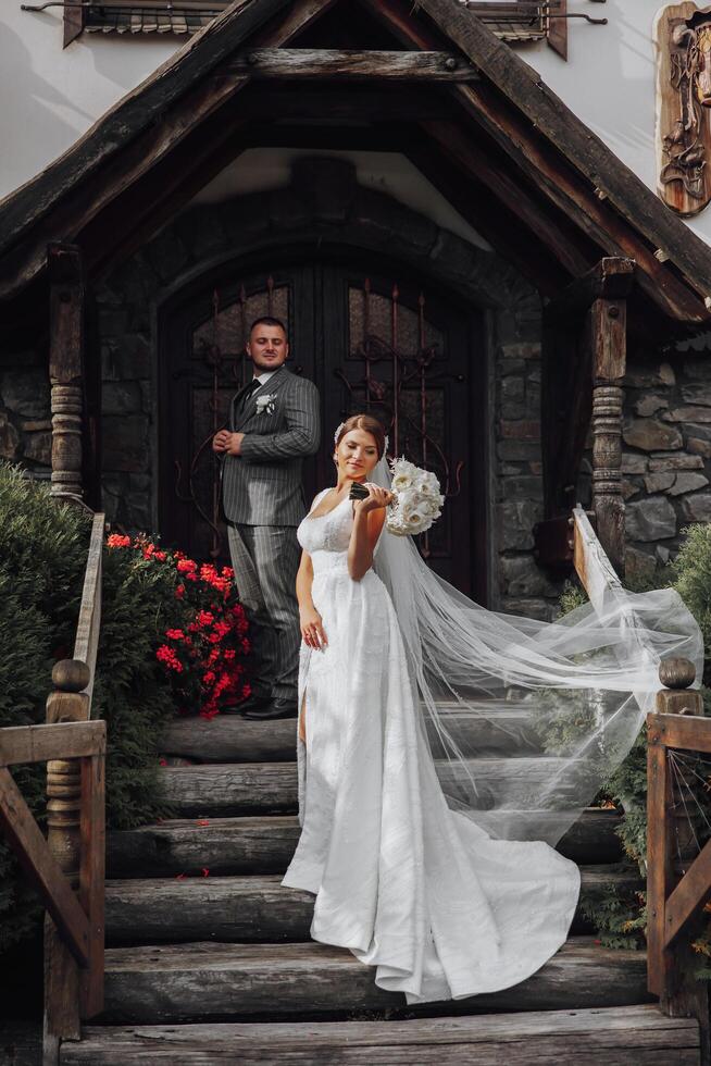 Romantic full-length portrait of a brown-haired woman in a white dress with a bouquet standing at the wooden gate of the entrance to the church, tattooed behind photo