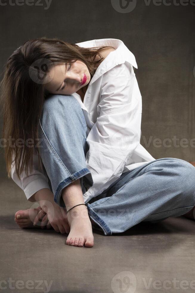 un niña en un blanco camisa y pantalones se sienta con su cabeza en su rodillas triste muchacha. foto