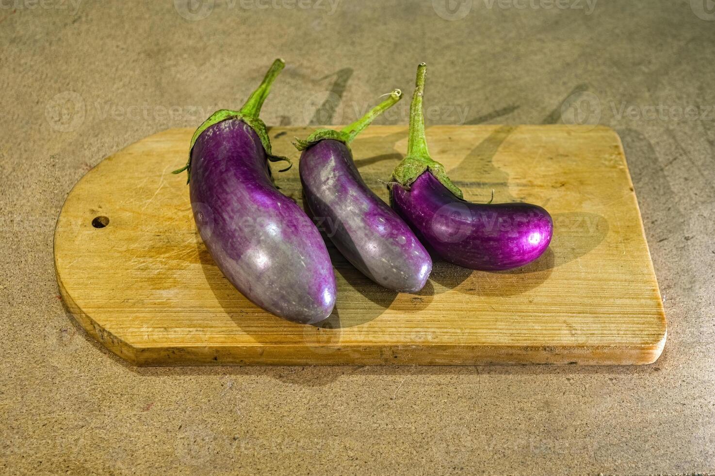 three purple eggplants or Solanum Melongena lying on a cutting board with a plain wooden board background photo