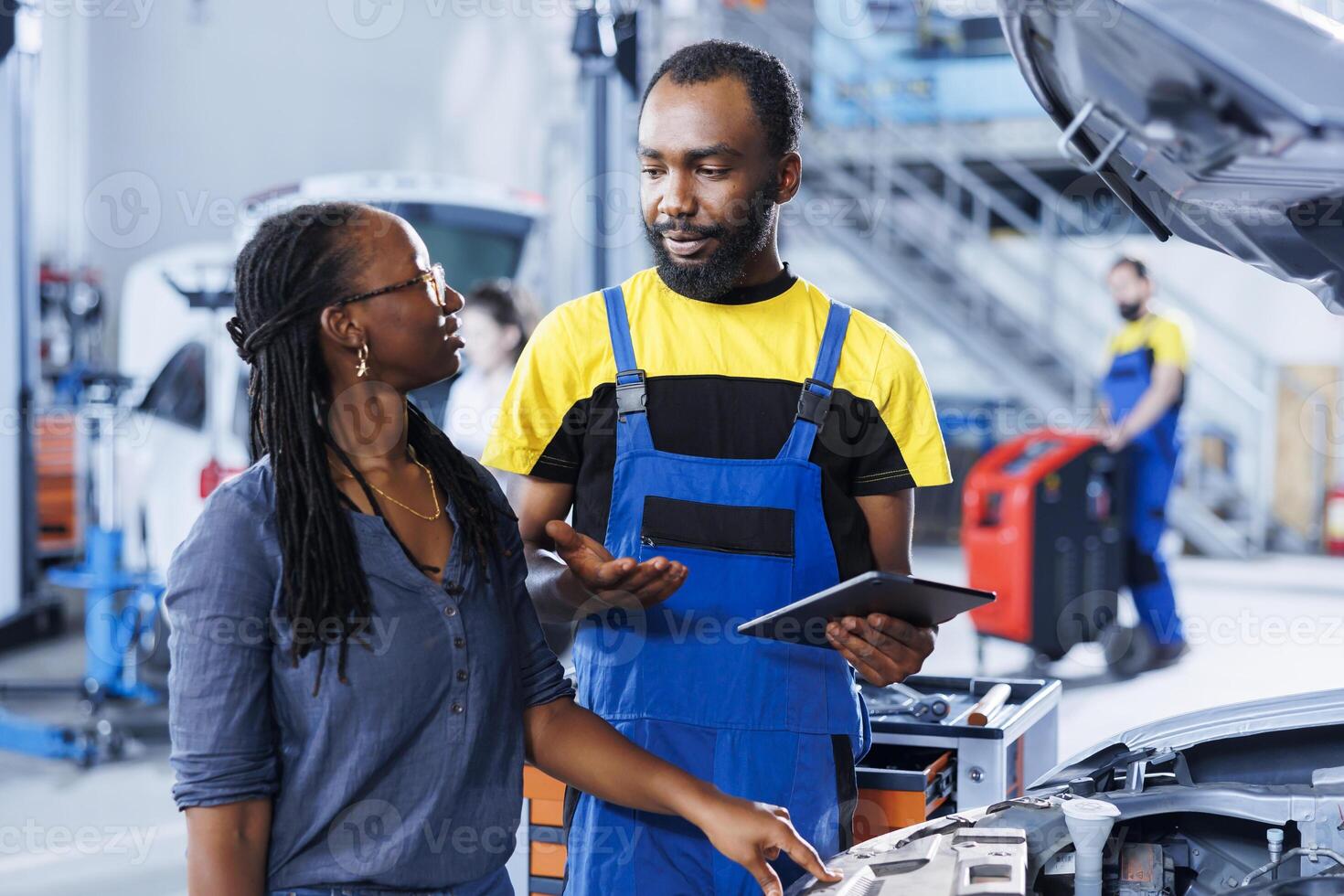 Mechanic at auto repair shop conducts annual vehicle checkup, informing customer about needed motor oil replacement. BIPOC garage worker talks with customer after finishing car inspection photo