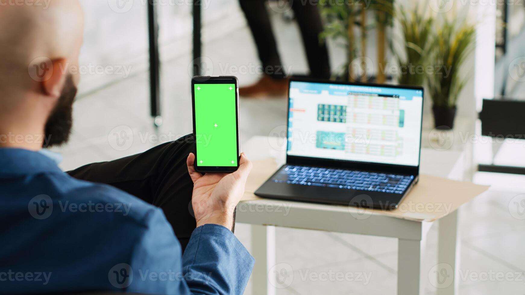 empleado sostiene teléfono con pantalla verde mientras él mira a ordenador portátil corriendo Estadísticas marcador. empresario con aislado monitor chromakey trabajando en compañía, exitoso agencia. foto