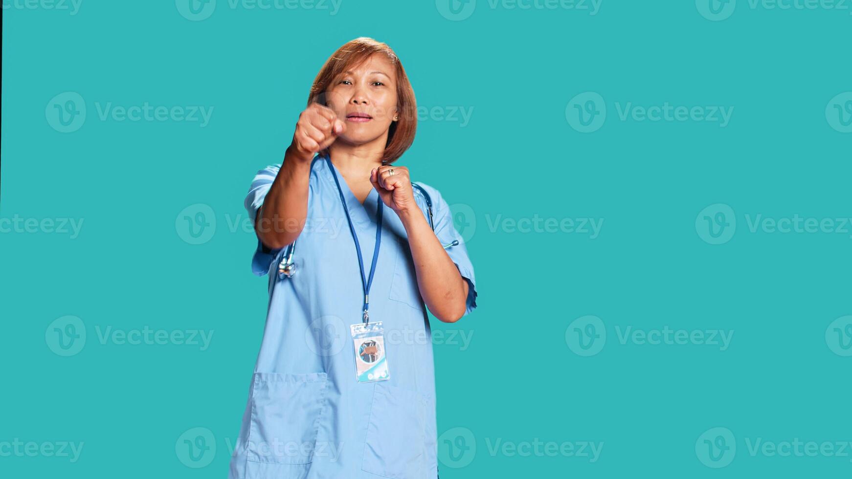 Happy playful asian nurse having fun pretending to fight, throwing punches. Funny high spirited healthcare specialist mock boxing while taking break from work, isolated over blue studio background photo