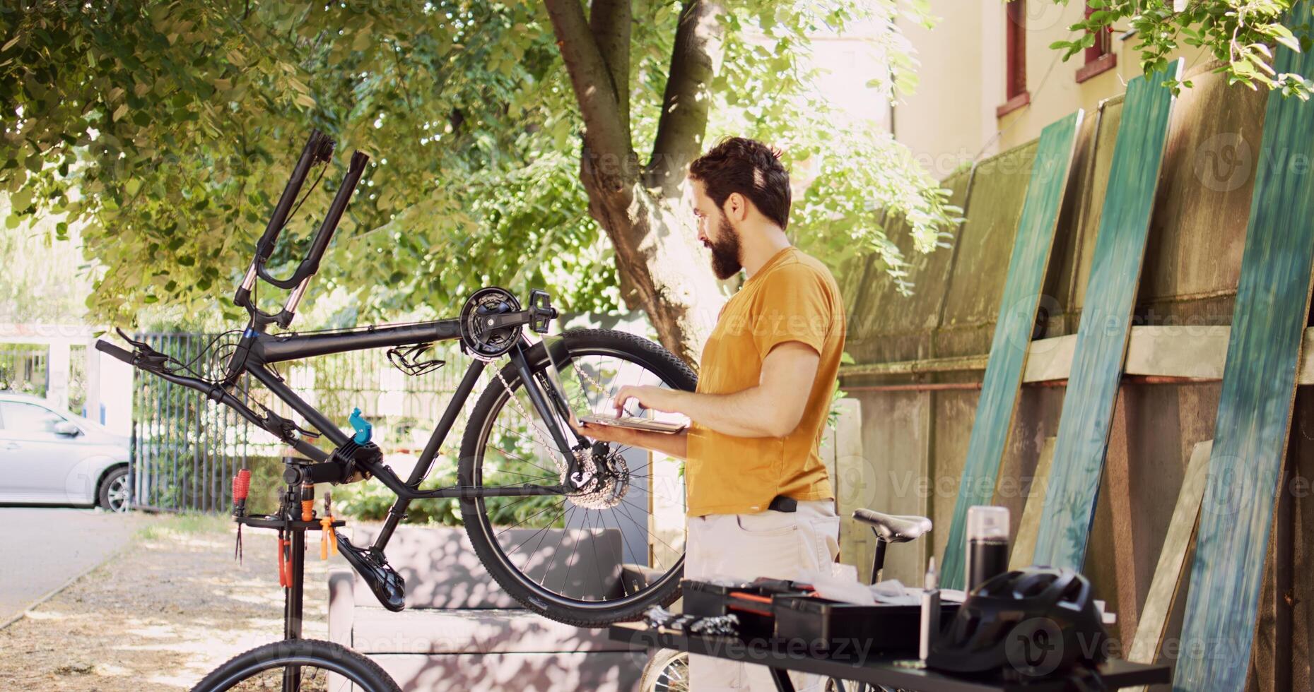 Male cyclist holding personal computer and inspects bike pedal and crank arm for damages in home yard. Active caucasian man enjoys browsing internet, fixing and maintaining modern bicycle. photo