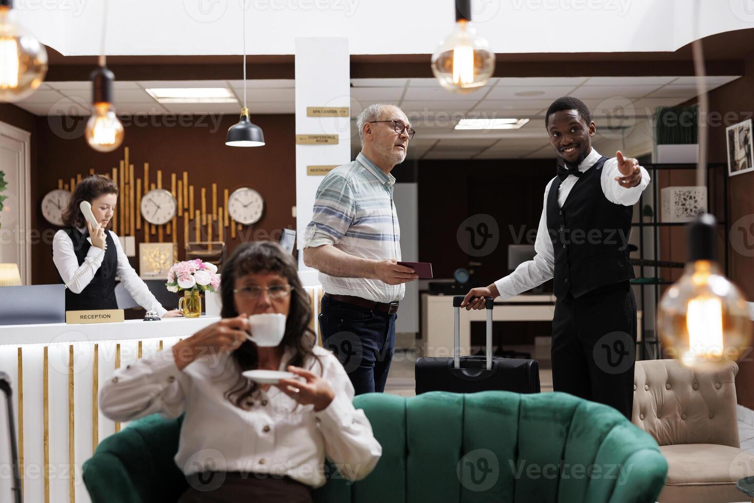 Friendly african american bellboy assists senior man in hotel reception with luggage to reserved room. Elderly female visitor sipping tea while waiting for check-in in comfy lounge area with sofas. photo