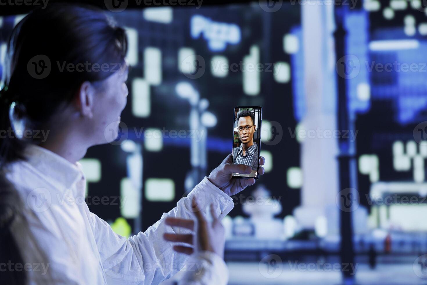 Asian man in videocall with step brother while walking around city avenues at night, giving him work updates. Person uses smartphone to show sibling dimly illuminated cityscape photo