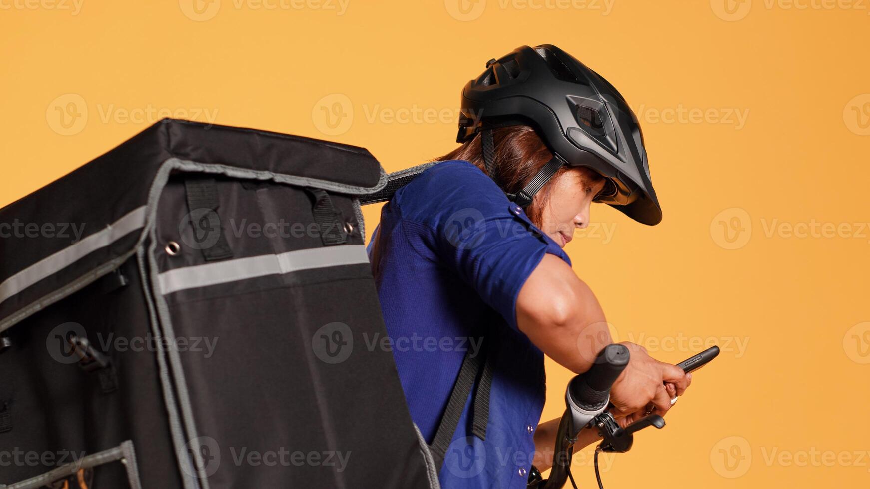 Courier imputing client order in phone app, preparing to ride bicycle to customer location. BIPOC cyclist isolated over orange studio background delivering takeout meals using thermal bag, close up photo