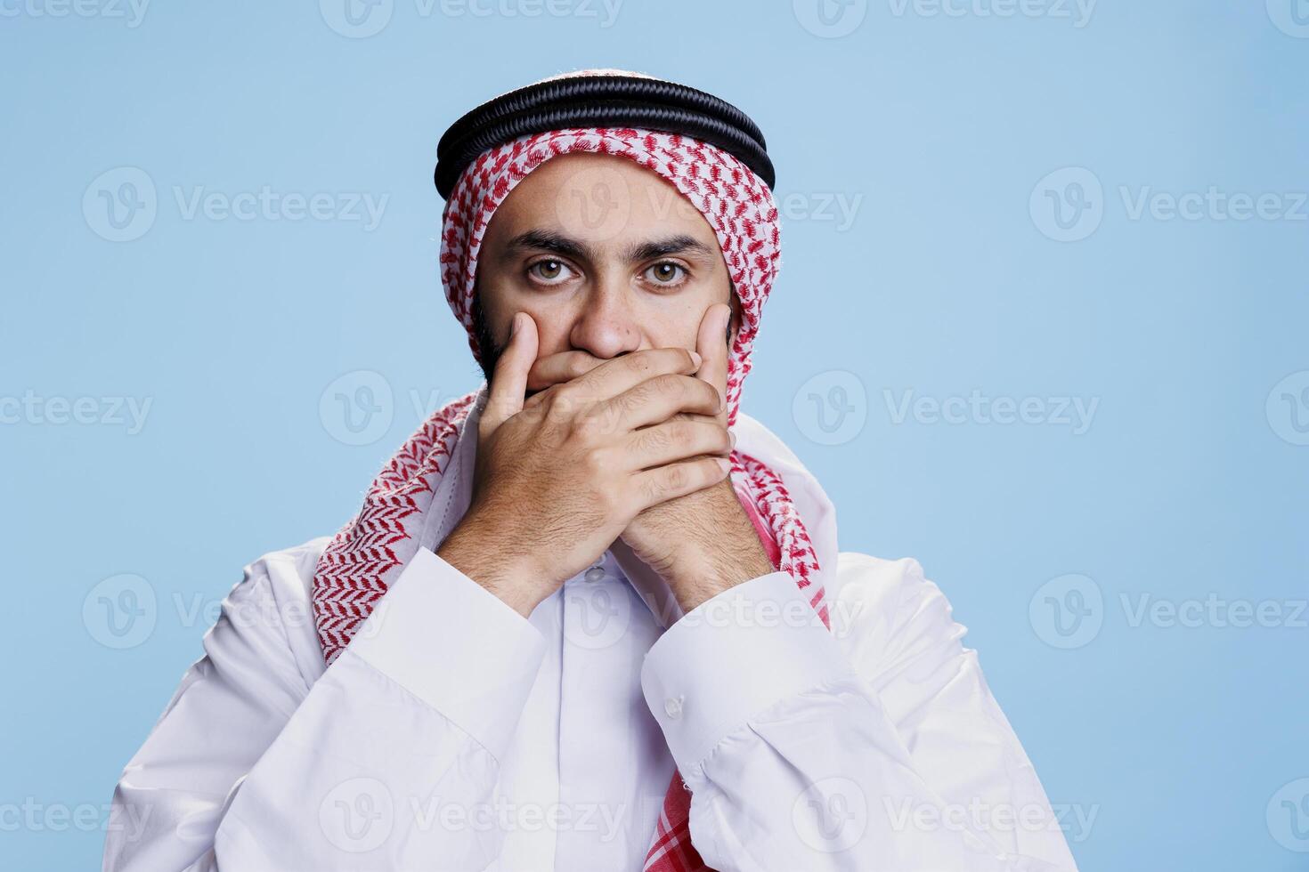 Man wearing traditional islamic clothes covering mouth with hands, showing speak no evil sign studio portrait. Muslim person keeping secret, showcasing three wise monkeys concept photo