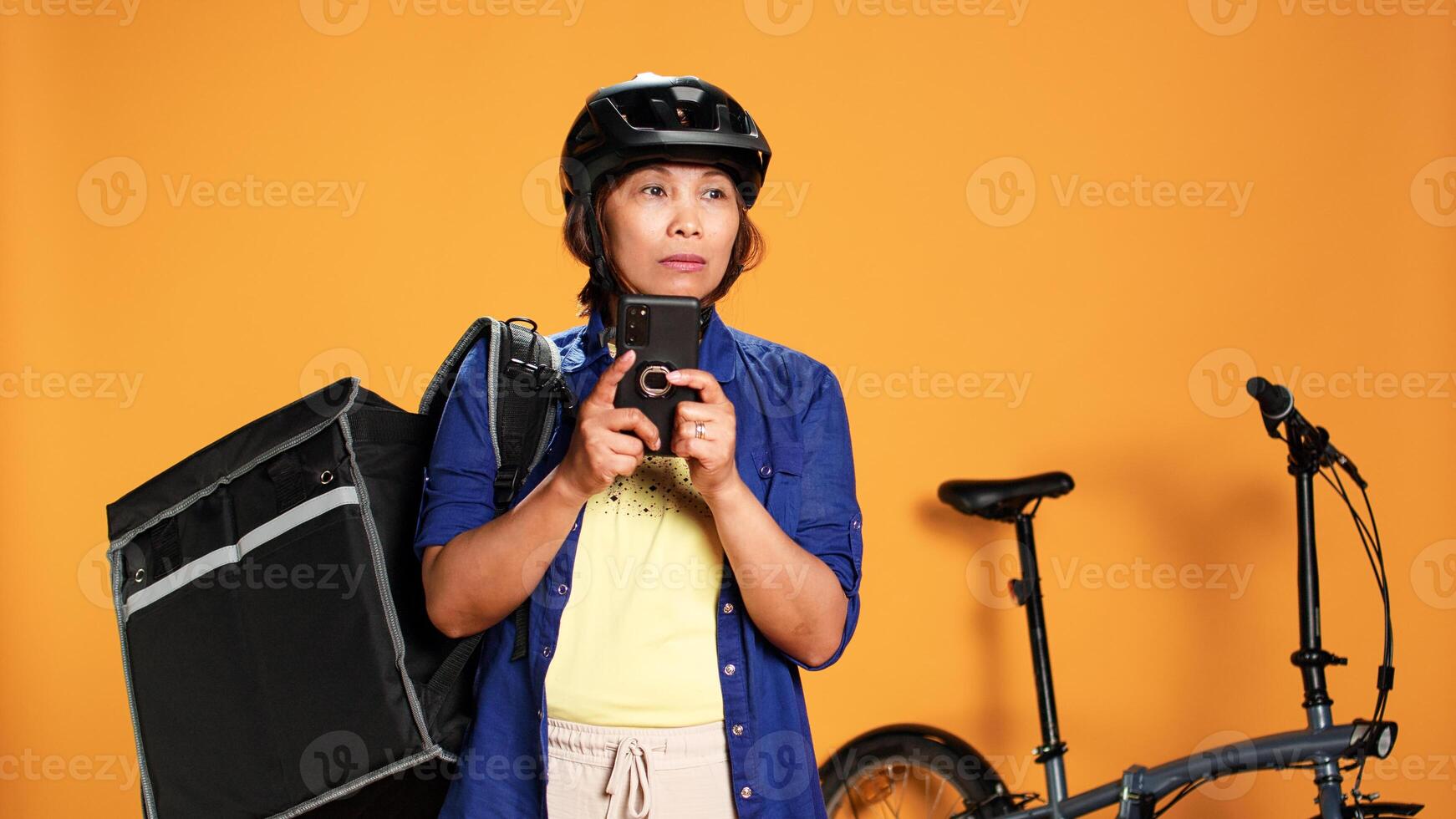 confuso asiático mujer mirando alrededor para direcciones, inseguro de cliente ubicación paradero. profesional mensajero vistiendo termico mochila montando bicicleta a entregar alimento, aislado terminado estudio antecedentes foto