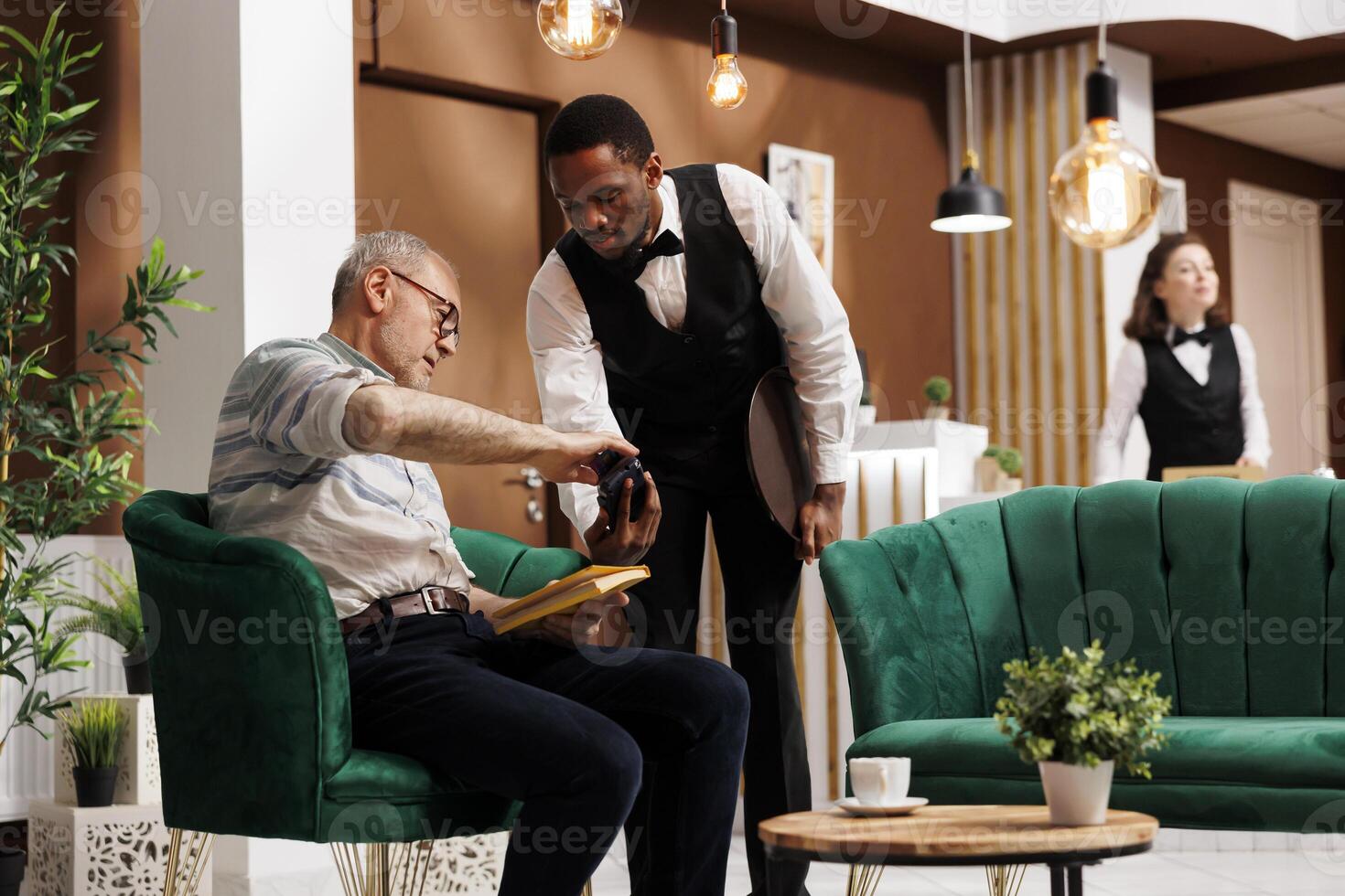 Retired old man pays with credit card at POS in hotel lobby, assuring quick and secure reservation transaction. Elderly guy with book makes cashless payment with help from african american waiter. photo