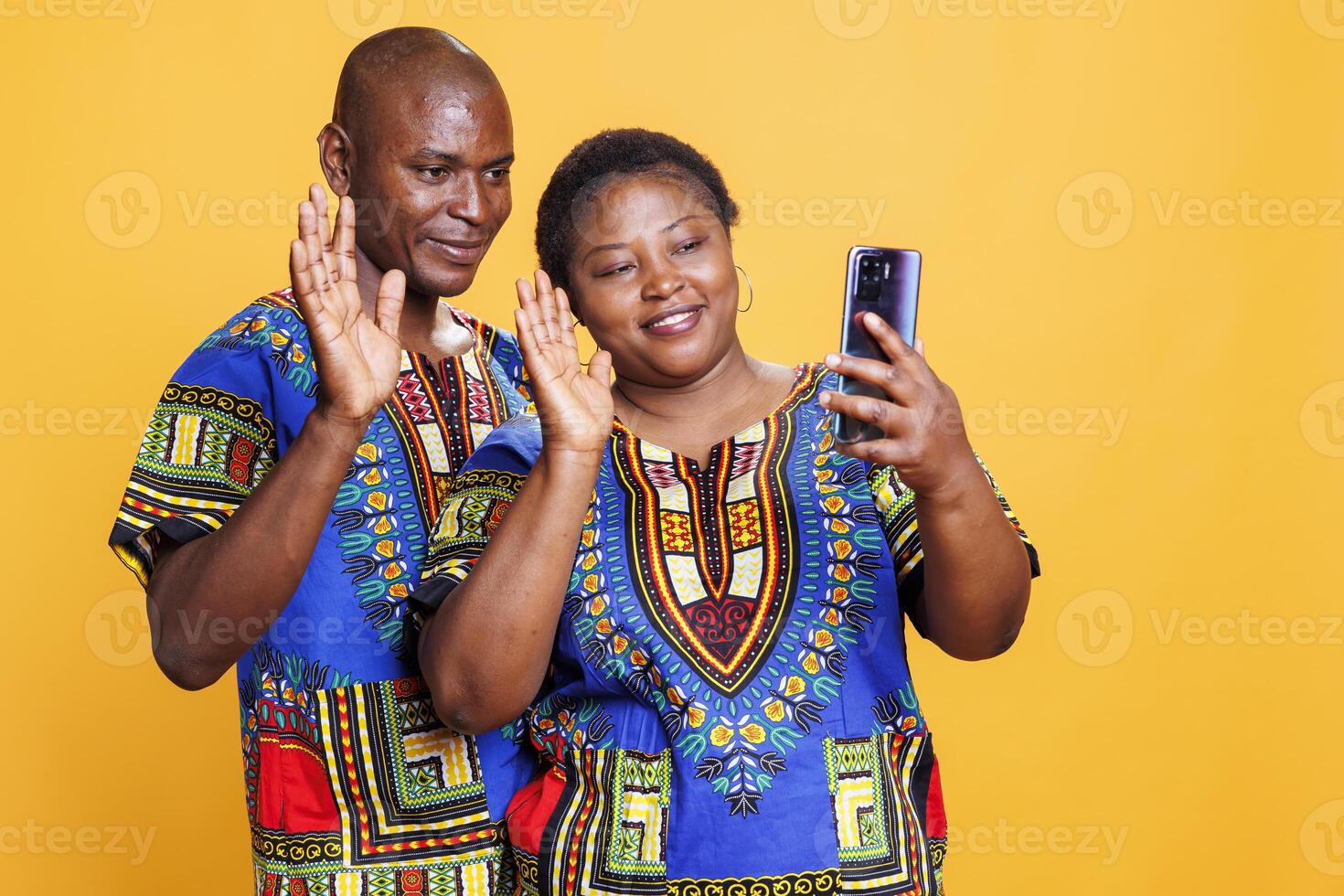 Smiling african american man and woman wearing ethnic clothes waving hi while speaking in smartphone videocall. Cheerful couple showing greeting gesture while having online communication on phone photo