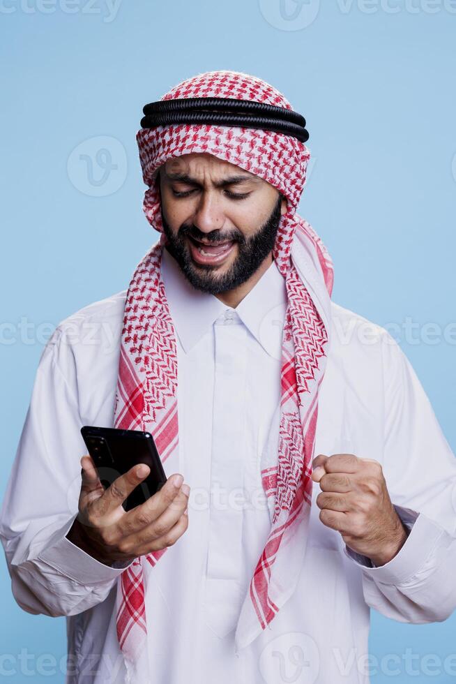 Excited man wearing traditional muslim outfit checking good news message while using smartphone. Happy cheerful muslim person showing winner gesture while scrolling internet page on mobile phone photo