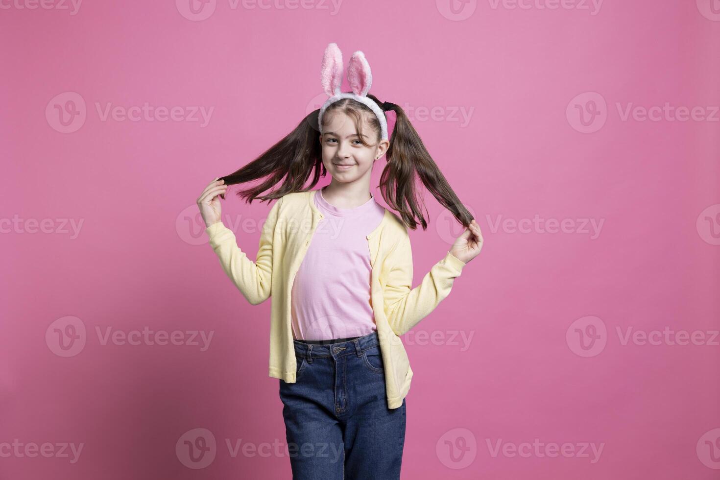 Cute small girl posing in studio with bunny ears and pigtails, celebrating easter and spring time. Adorable cheerful child standing over pink background, seasonal traditions and festive toys. photo