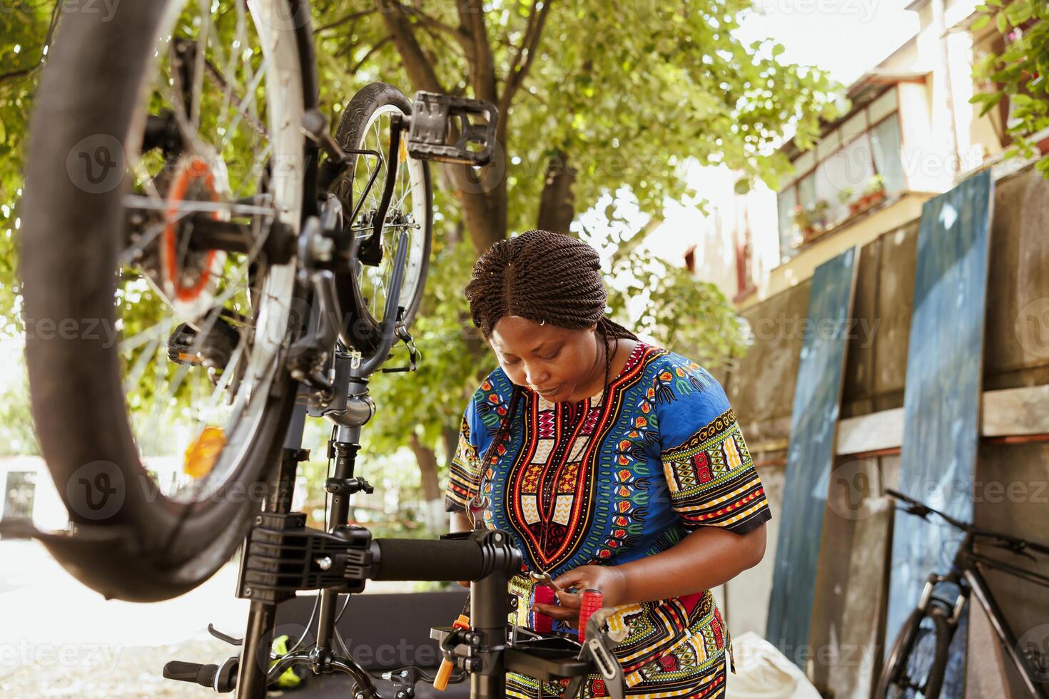 Energetic african amercian female cyclist carefully inspecting specialized equipment for servicing bicycle outside. Sports-loving black woman searching for work tool in home yard dedicated to summer bike maintenance. photo