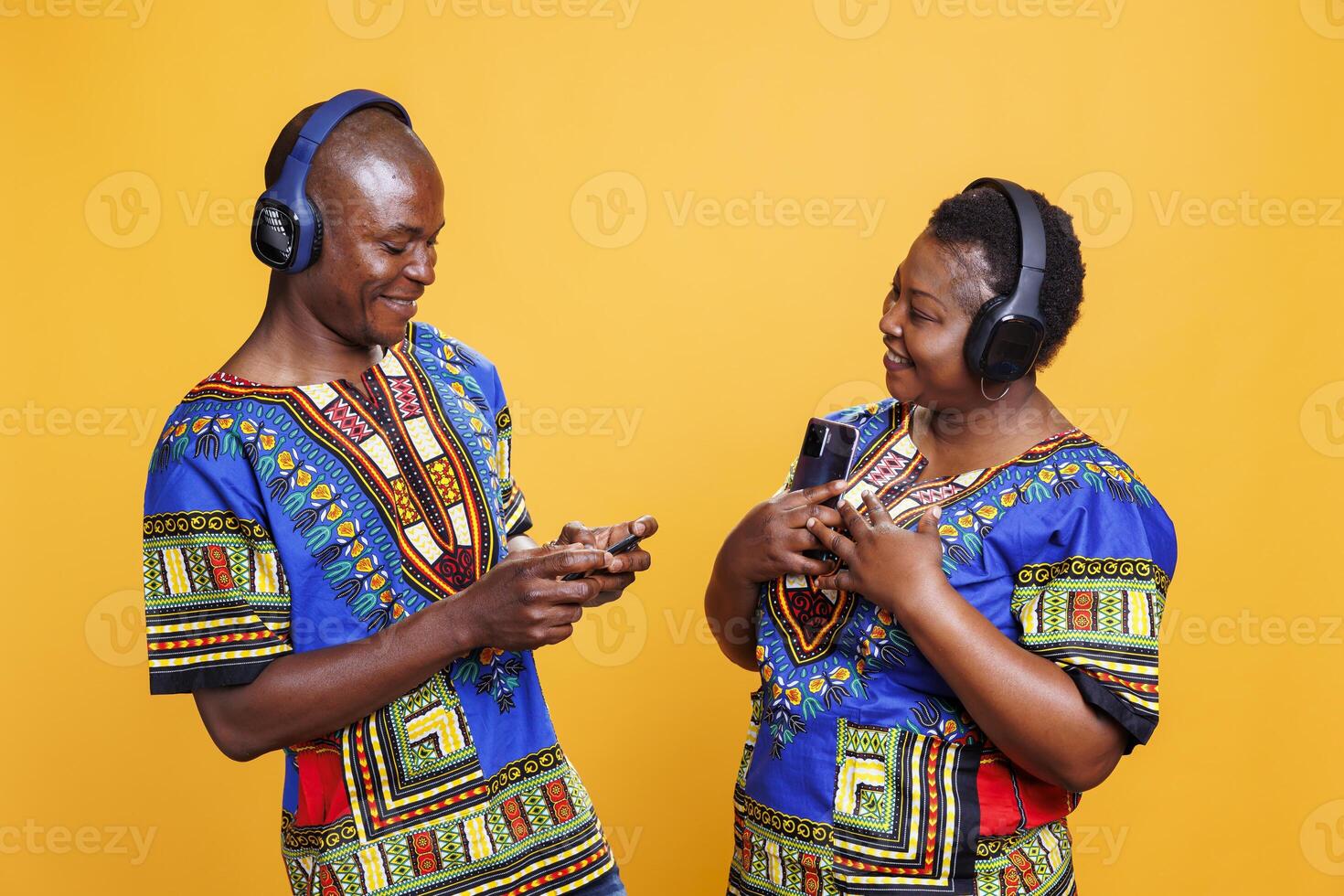 Smiling black man and woman wearing ethnic clothes listening to music in headphones. Cheerful couple in relationship holding mobile phones and enjoying playlist in wireless earphones photo