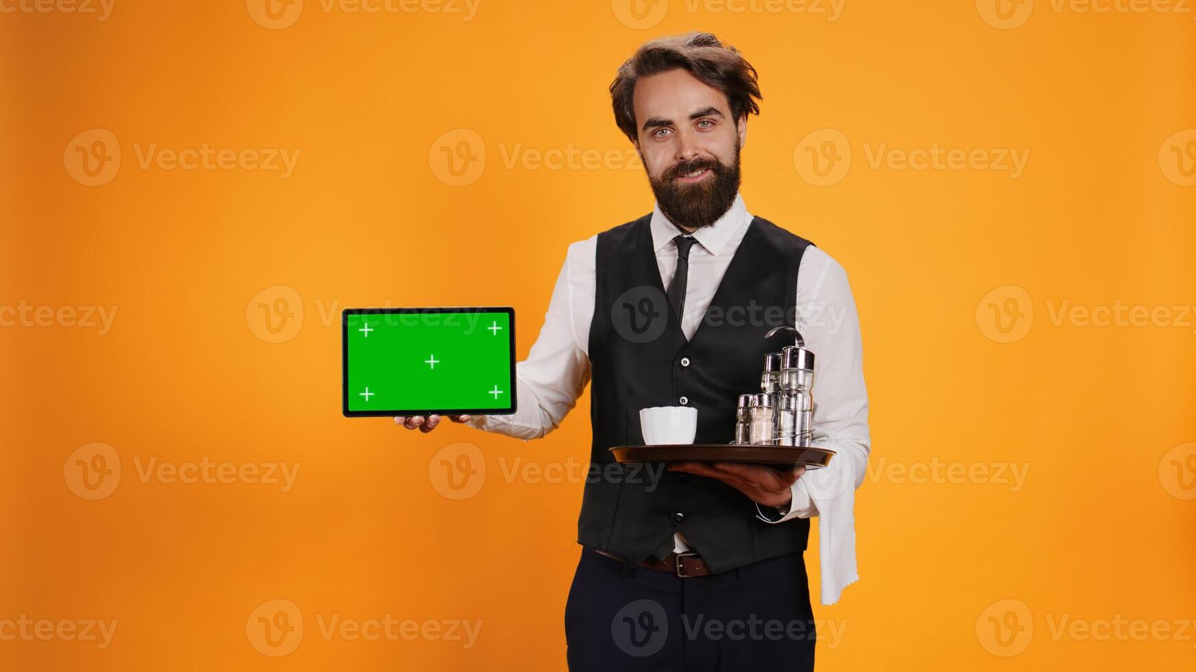 Joyful employee shows greenscreen on tablet, posing with food platter against yellow background. Five star waiter presenting blank isolated mockup template on display in studio, formal attire. photo