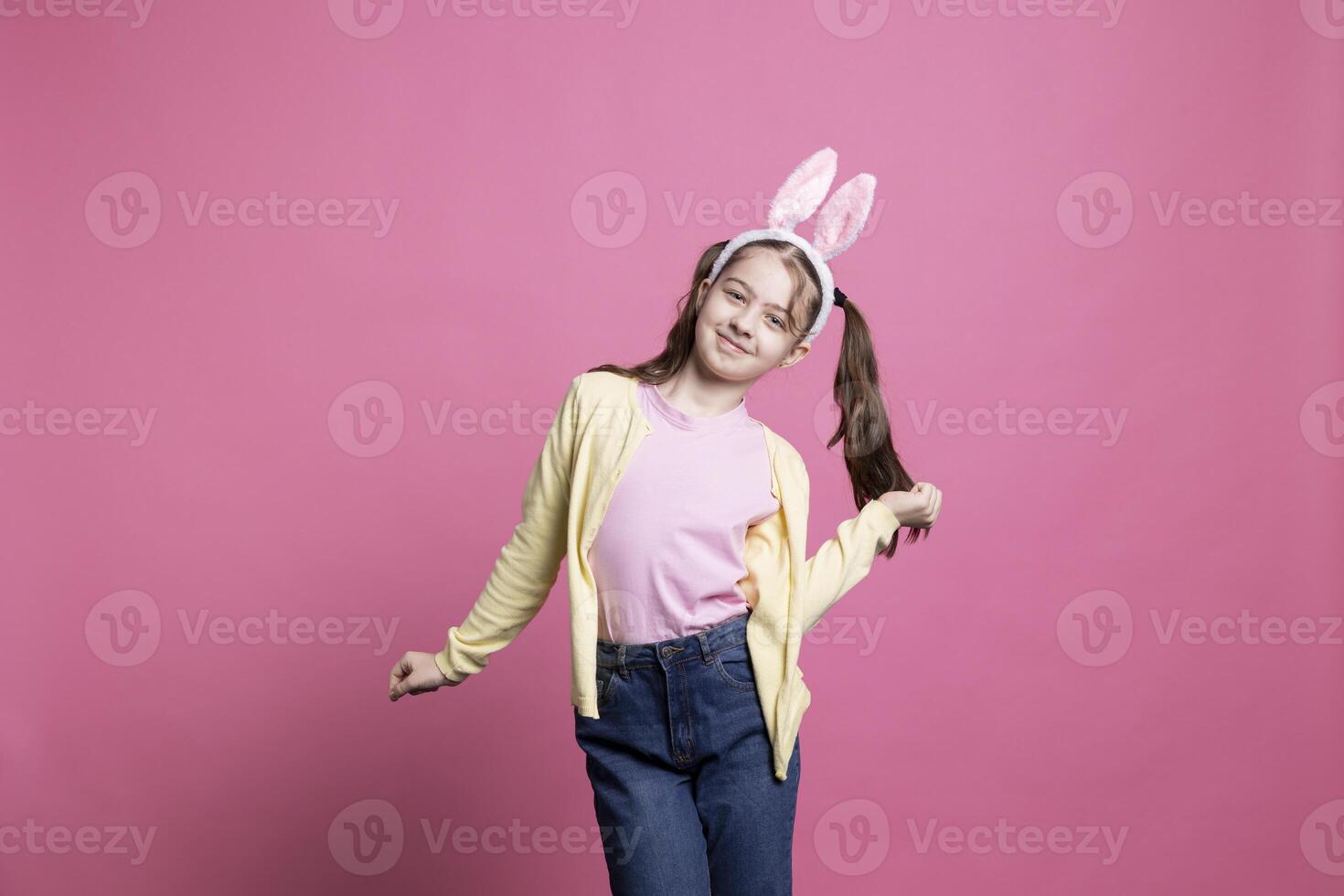 Cheery little girl dancing and having fun in front of camera, feeling happy and excited about easter celebration. Confident small toddler showing dance moves while she wears bunny ears. photo