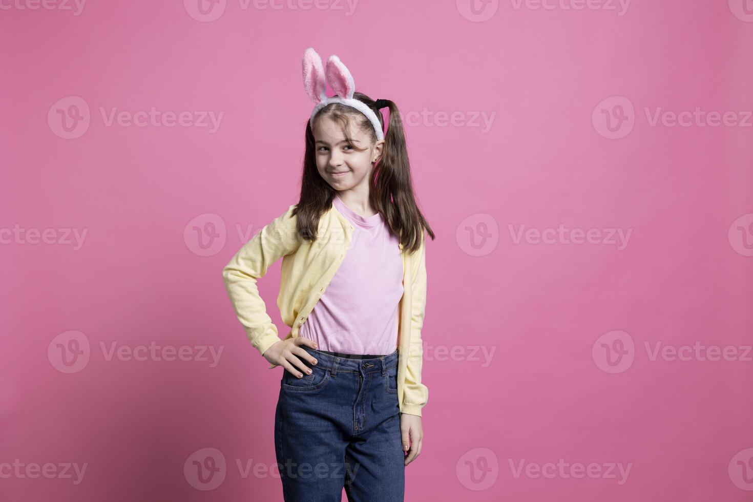 Sweet child wearing bunny ears posing in front of the camera, standing against pink background. Young girl feeling happy and joyful, adorable kid celebrating easter time in studio. photo
