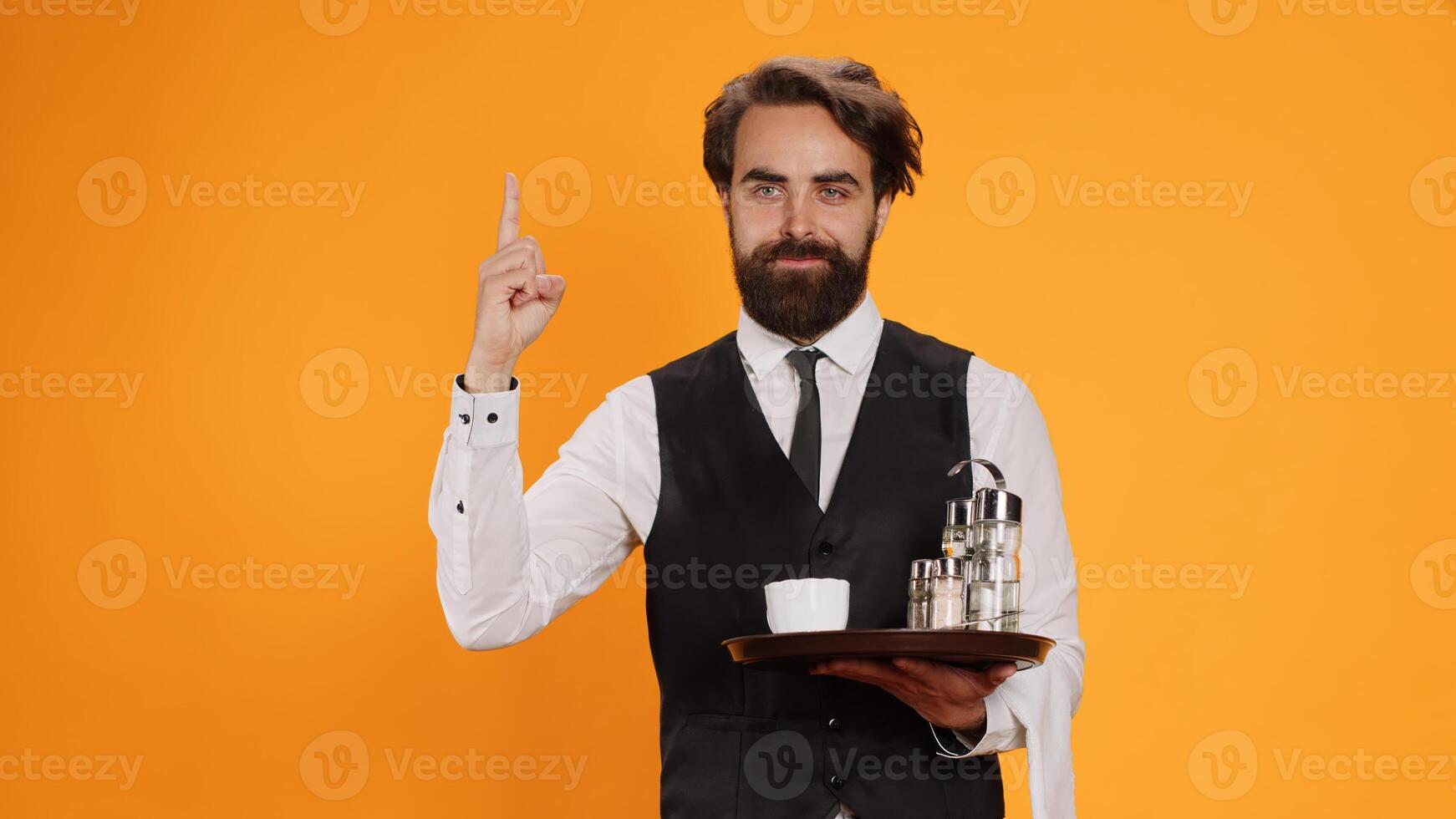 Elegant butler having new idea on camera, raising index finger and indicating upwards direction with restaurant food tray in hand. Skilled waiter feeling genious, innovative with fresh thoughts. photo