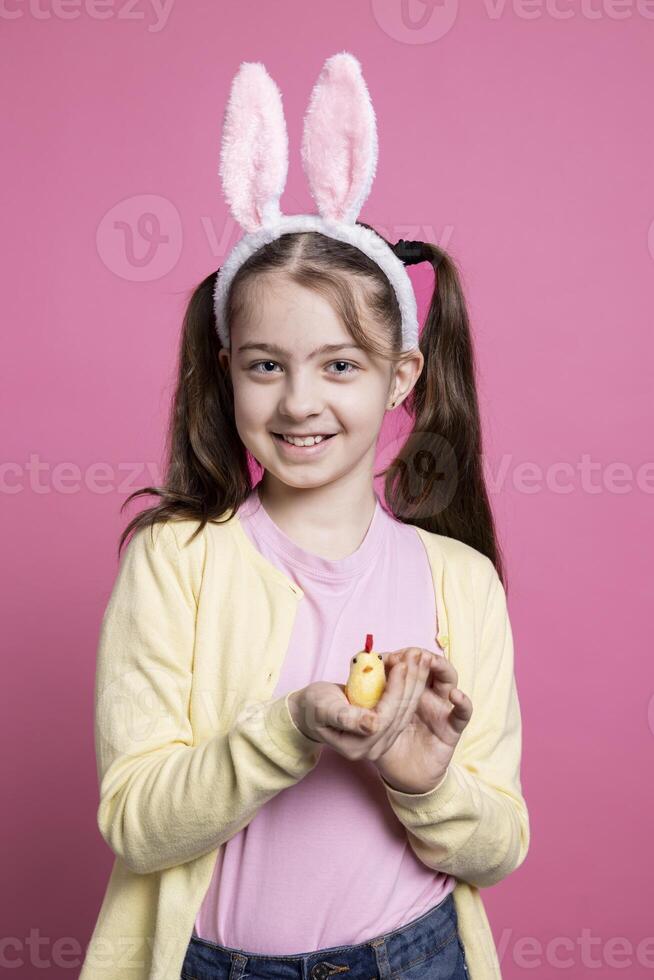 Little smiling girl holding an adorable stuffed chick in front of camera, cheerful young kid with fluffy golden toy over pink background. Small child with cute bunny ears for festivity. photo