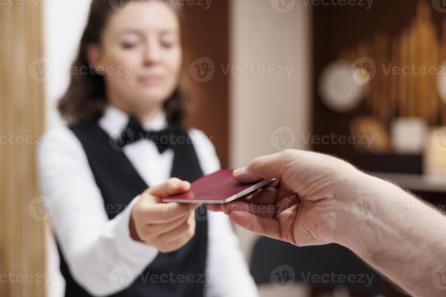 Female receptionist helps elderly man at front desk with check in and confirm bookings. Close-up of retired senior male tourist giving personal document to the caucasian concierge for verification. photo