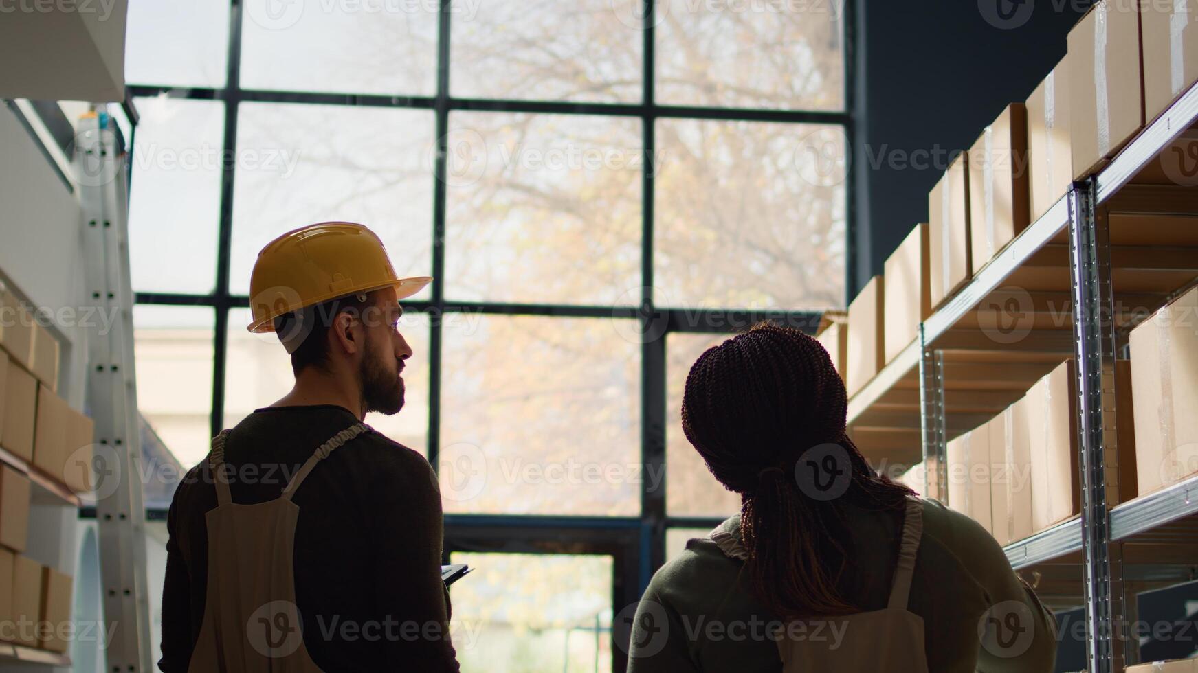 Meticulous trainee walking through warehouse rows scanning cardboard boxes on shelves under team leader guidance, using digital devices to update info before sending parcels for delivery photo