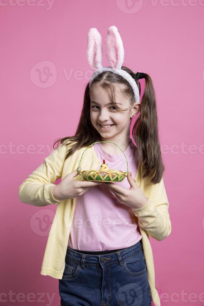Lovely little kid with rabbit ears poses with adorable springtime toys while carrying an Easter basket full of eggs and a chick. Joyful and enthusiastic youngster enjoying the holidays in studio. photo