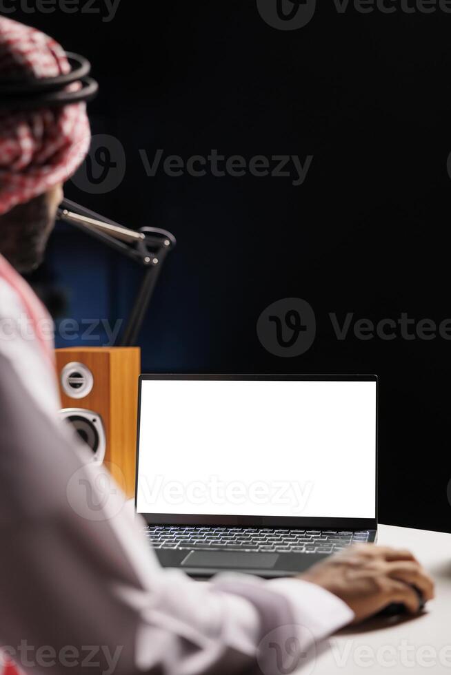 Image depicts Muslim guy working from home, sat at his desk, with an isolated chromakey template shown on his minicomputer. Young Arab man using laptop with a copyspace white screen. photo