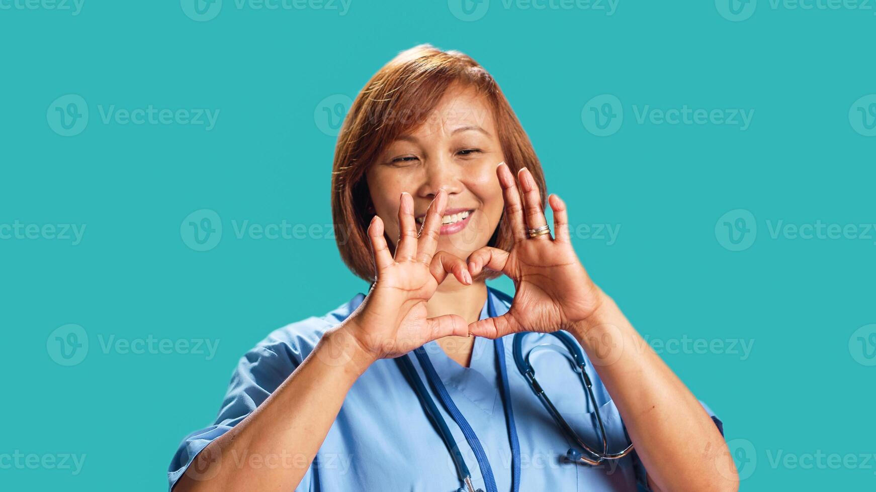 Close up shot of joyful qualified BIPOC nurse doing heart symbol shape with hands. Clinic employee showing love gesturing while at work, isolated over blue studio background photo