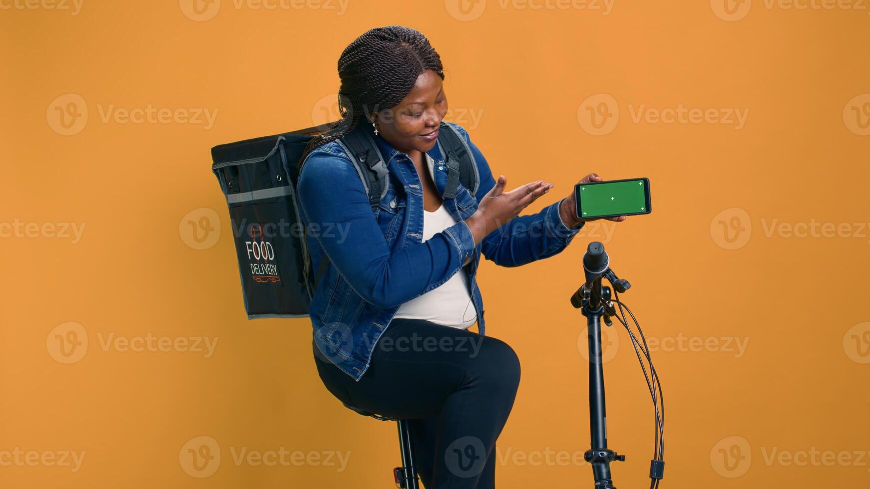 African american courier gripping cell phone showing green screen display while riding bicycle. Black woman demonstrates cutting-edge delivery app on mobile device with isolated mockup template. photo