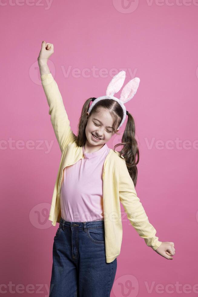 Happy toddler with pigtails and fluffy ears fooling around in the studio, playing and acting silly in front of the camera. Joyful little girl dancing with confidence over pink background. photo