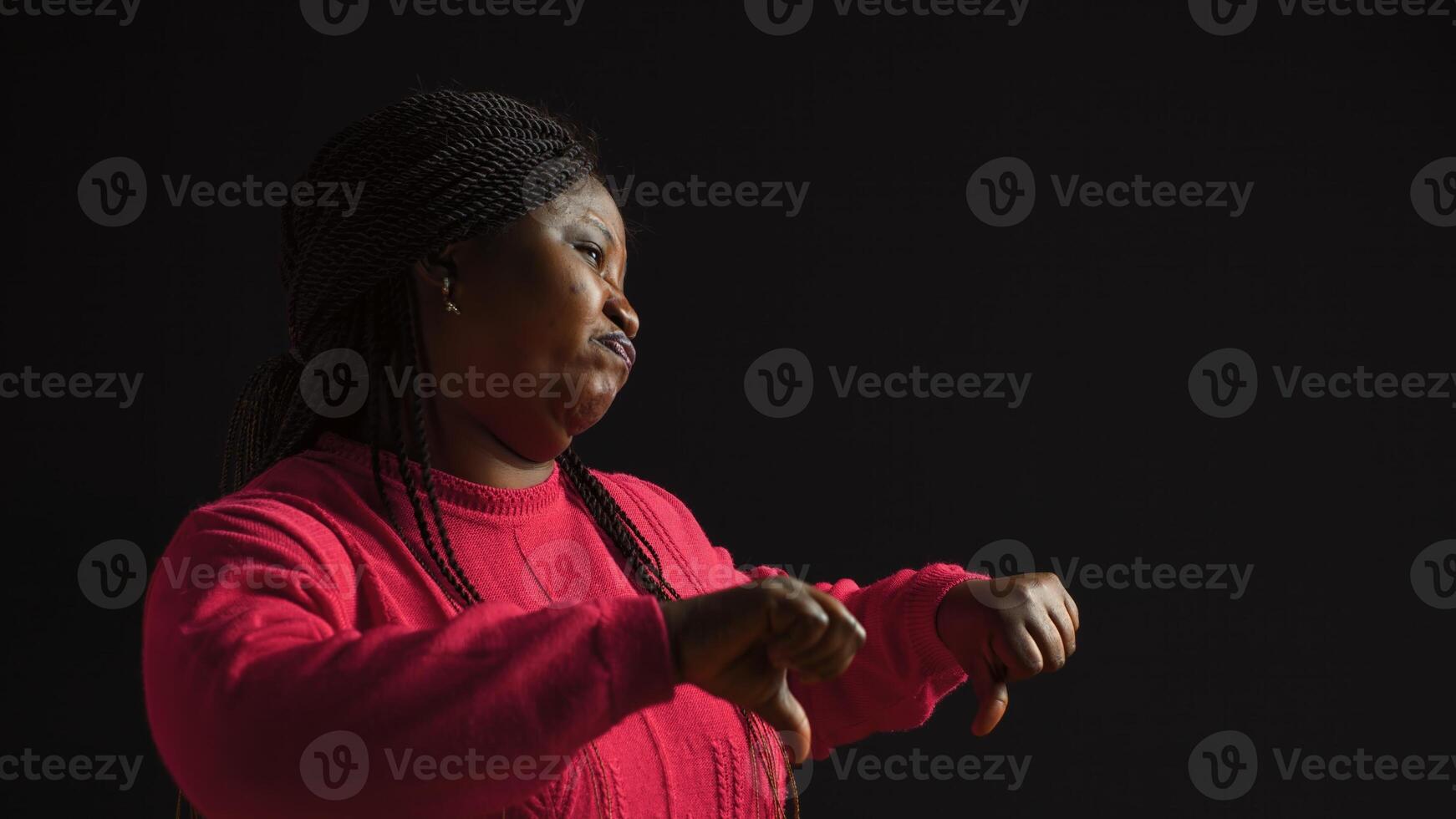 Standing against black background fashion model shows disapproval by holding her hands in front of her. African American woman in pink sweater uses thumbs down motion to disagree. Side-view shot. photo