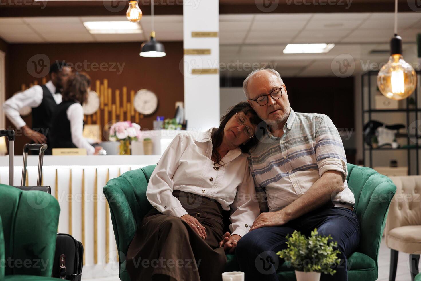 Exhausted beautiful elderly couple sleep on comfortable couches in luxurious hotel lobby. Waiting for check-in, retired senior travelers taking a nap in lounge area after a long flight. photo