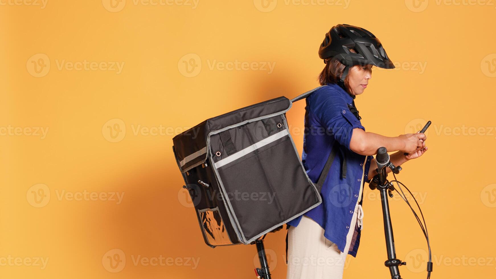 Mano Disparo de bipoc ciclista entregando eliminar comida a clientela, mecanografía siguiente habla a en teléfono aplicación mensajero montando bicicleta, vistiendo protector engranaje y térmico bolsa, aislado terminado estudio antecedentes foto